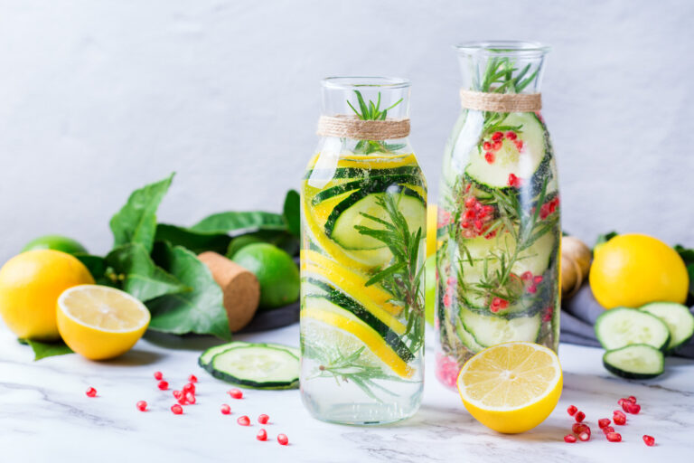 Zwei Glaskaraffen mit Wasser, Gurken, Zitronen und Kräutern, umgeben von frischen Zutaten auf einer Marmorfläche.
